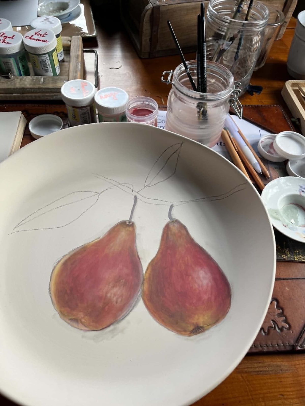 Une assiette en céramique en cours de peinture à La Roseraie : deux poires déjà peintes, la branche encore au crayon, entourée des pots de couleur et des pinceaux de Carine