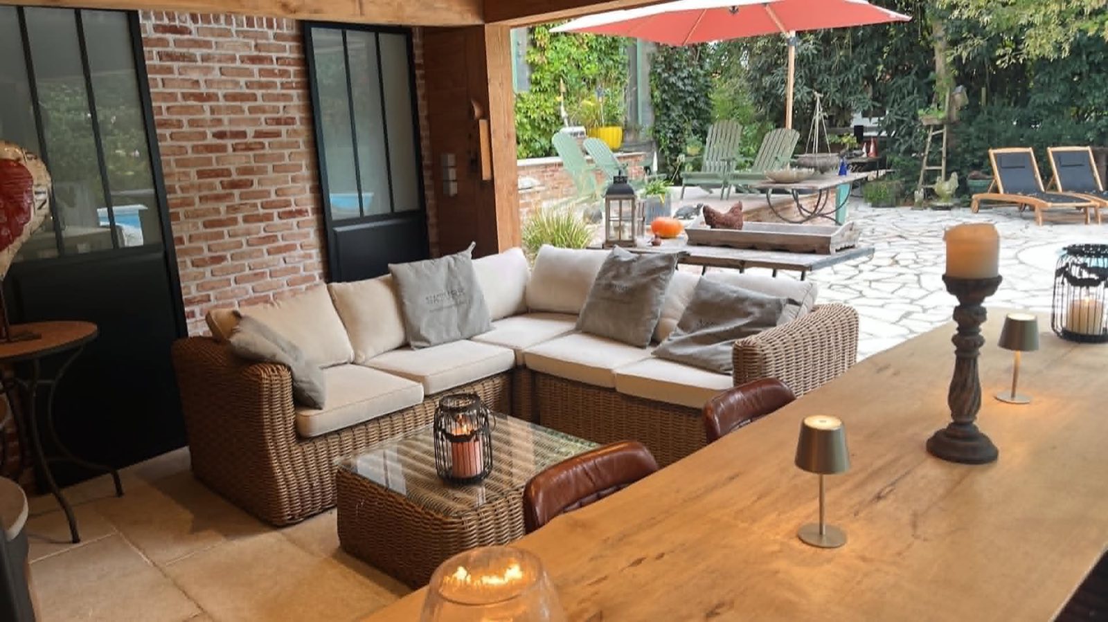 L'intérieur de la cuisine d'été de La Roseraie : mur de brique, verrière, salon en rotin, et la terrasse de la piscine au fond