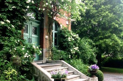 Le perron de la maison de maître de La Roseraie : la double porte verte, les marches de pierre et les urnes fleuries, sous les rosiers grimpants blancs