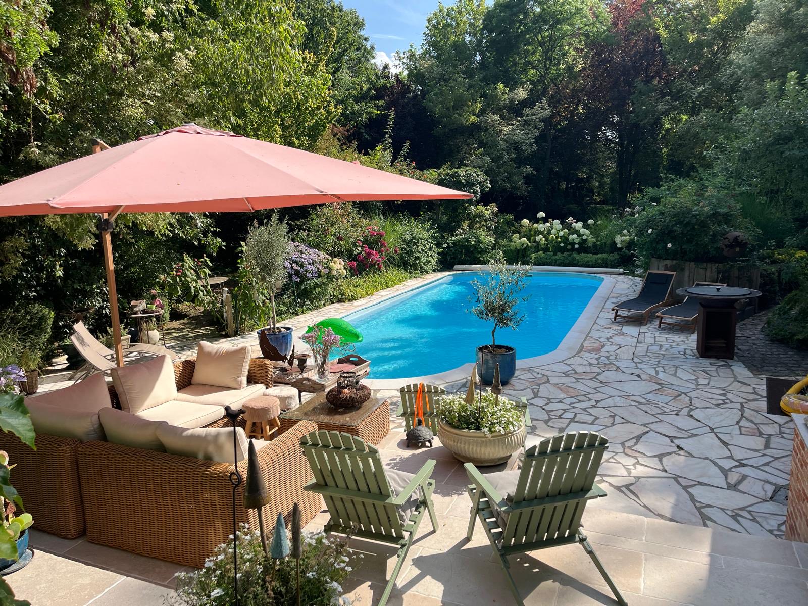 La terrasse et la piscine chauffée de La Roseraie en plein été : le salon en rotin sous le grand parasol, les chaises de jardin, les transats au bord de l'eau, un olivier en pot et les hortensias en fleurs